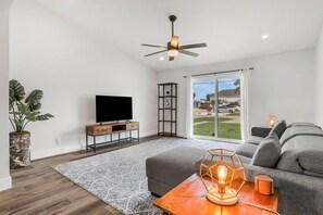 Ein modernes Wohnzimmer mit Flachbildfernseher, Deckenventilator, großem Fenster mit Aussicht, grauem Sofa und einer dekorativen Tischleuchte.