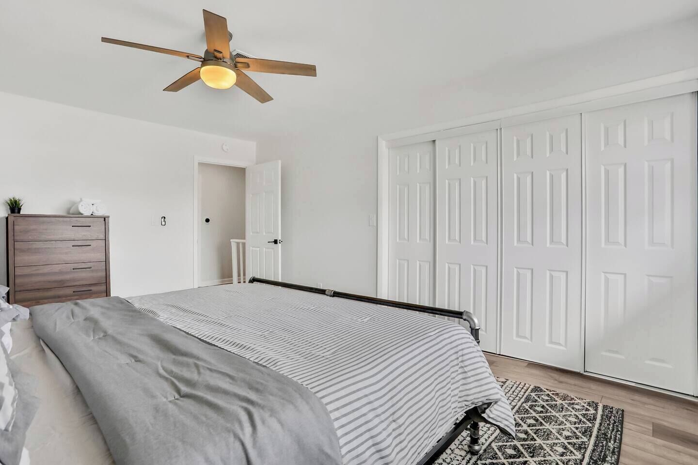 Une chambre à coucher avec un lit, une commode en bois, un ventilateur de plafond et une porte double blanche.