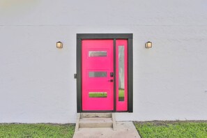 Eine leuchtend rosa Tür mit schwarzem Rahmen, in eine weiße Wand eingelassen, mit zwei Wandleuchten.