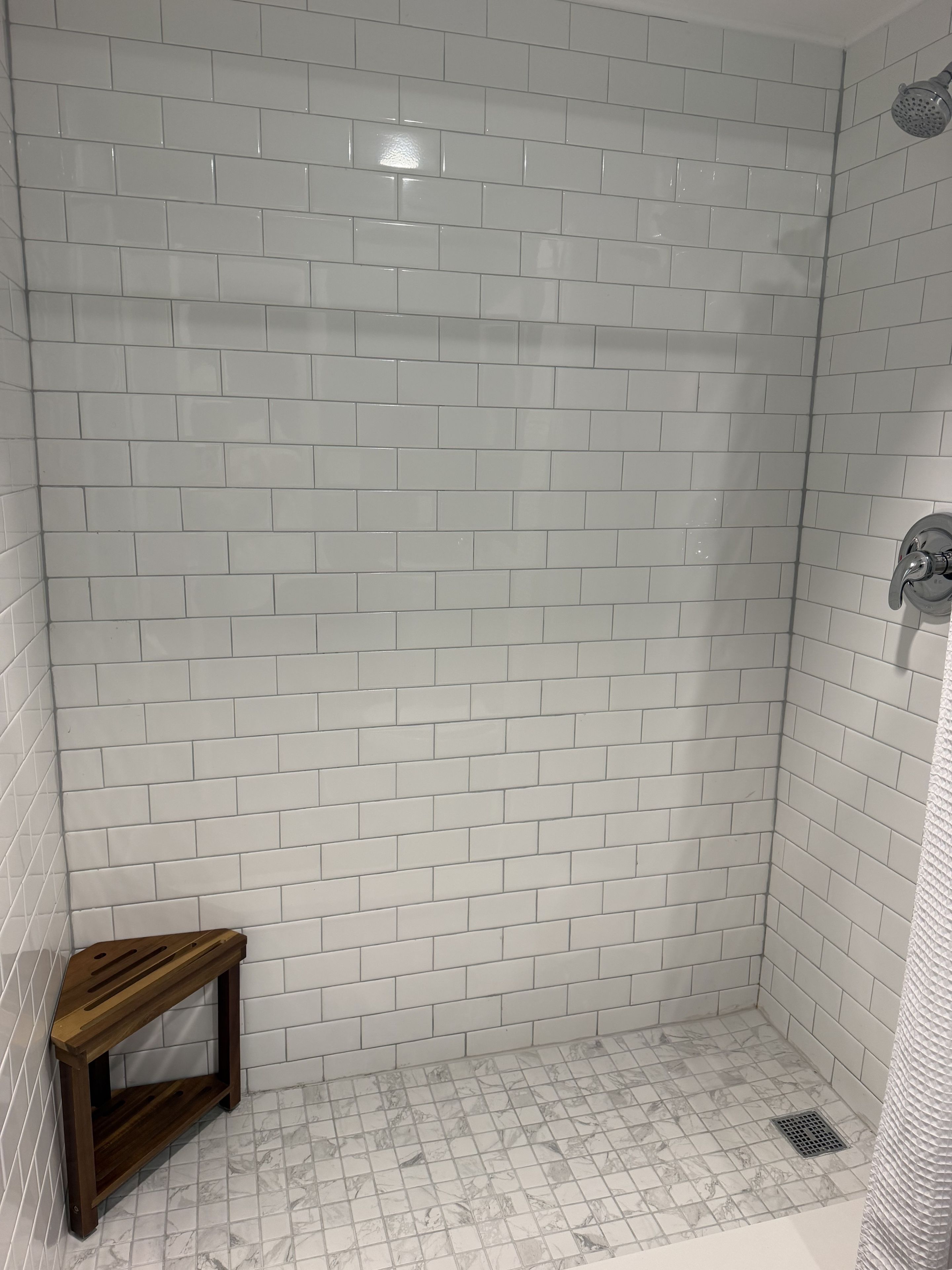 A shower area with white tiled walls and floor, a wooden bench, and a showerhead.