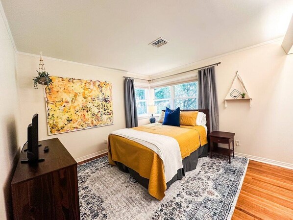 A bedroom with a bed, a television on a stand, a window with curtains, and a wall-mounted shelf.