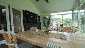 Uma sala de jantar com mesa de madeira, cadeiras de vime, televisão presa a uma parede de tijolos e vista através de grandes janelas.