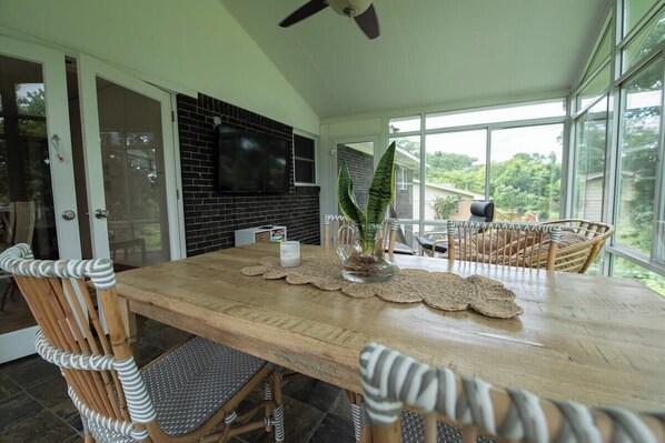 A dining area with a wooden table, wicker chairs, a television mounted on a brick wall, and a view through large windows.