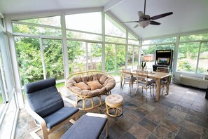 A sunroom with large windows, a ceiling fan, a dining table with chairs, and a gaming console.