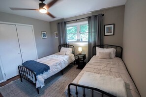A bedroom with two single beds, a ceiling fan, a window with curtains, and a bedside table with a lamp.
