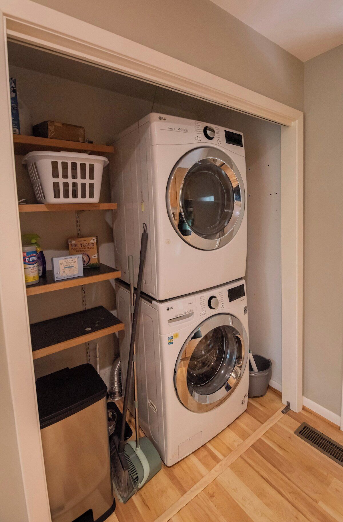 A laundry room with stacked washers and dryers, a broom, a trash can, and shelves with laundry supplies.