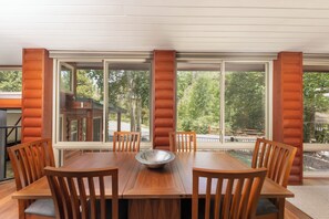 A dining area with a wooden table and chairs, large windows, and a view of trees.
