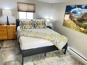 A bedroom with a bed, a nightstand, a lamp, a window with blinds, and a painting of a mountain landscape.