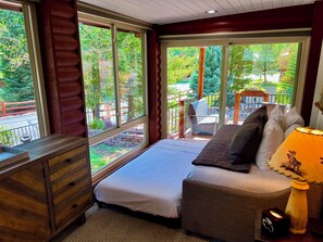 A bedroom with a bed, a wooden dresser, and a window with a view of a balcony and greenery.