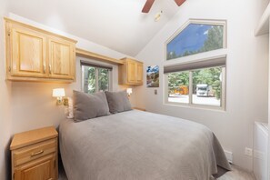 Un dormitorio con cama, armarios de madera, una mesita de noche, dos ventanas y vistas al exterior.