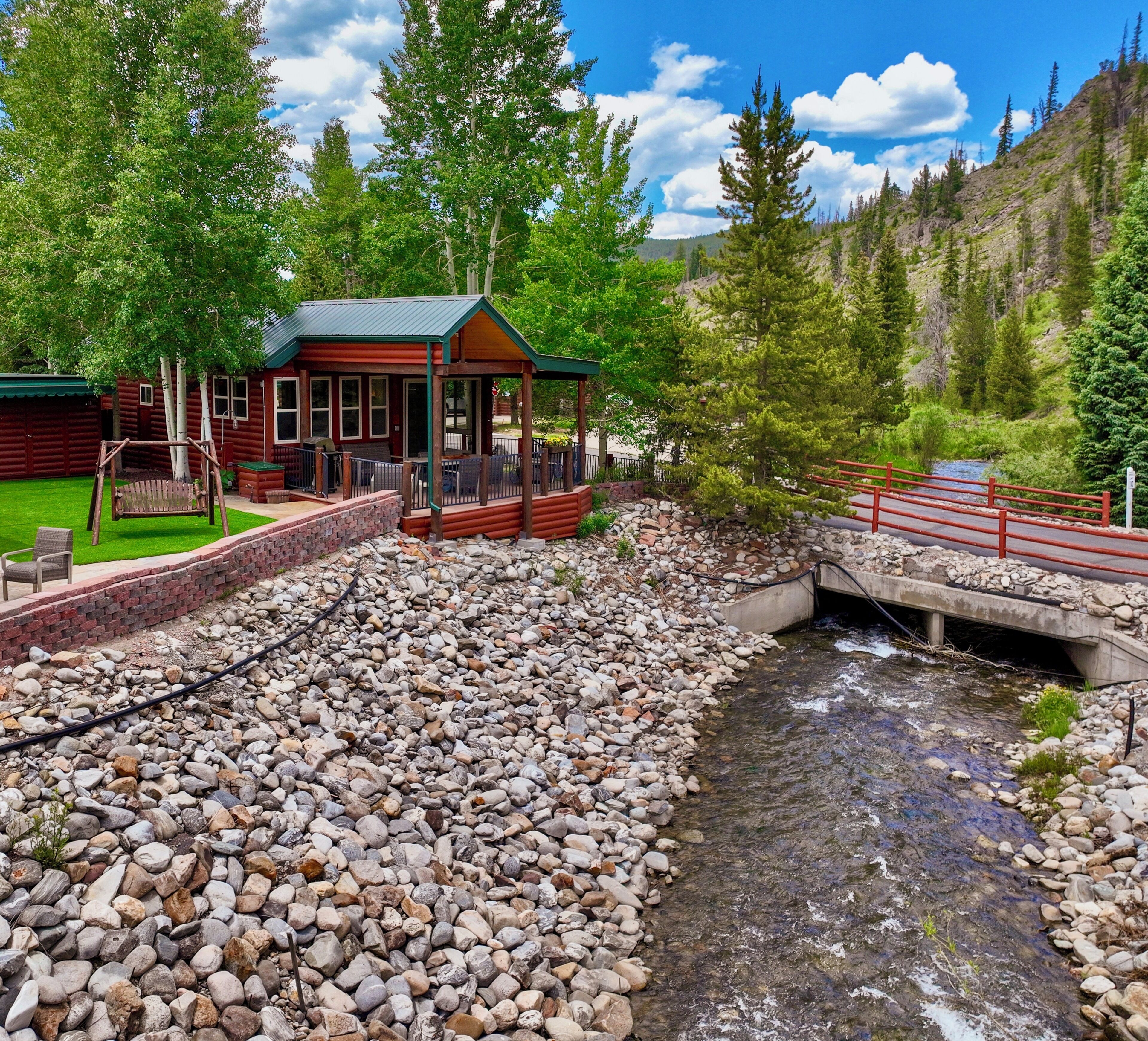 A log cabin with a porch and a swing, a small bridge over a stream, and a forested hillside in the background.