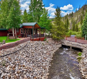 A log cabin with a porch and a swing, a small bridge over a stream, and a forested hillside in the background.