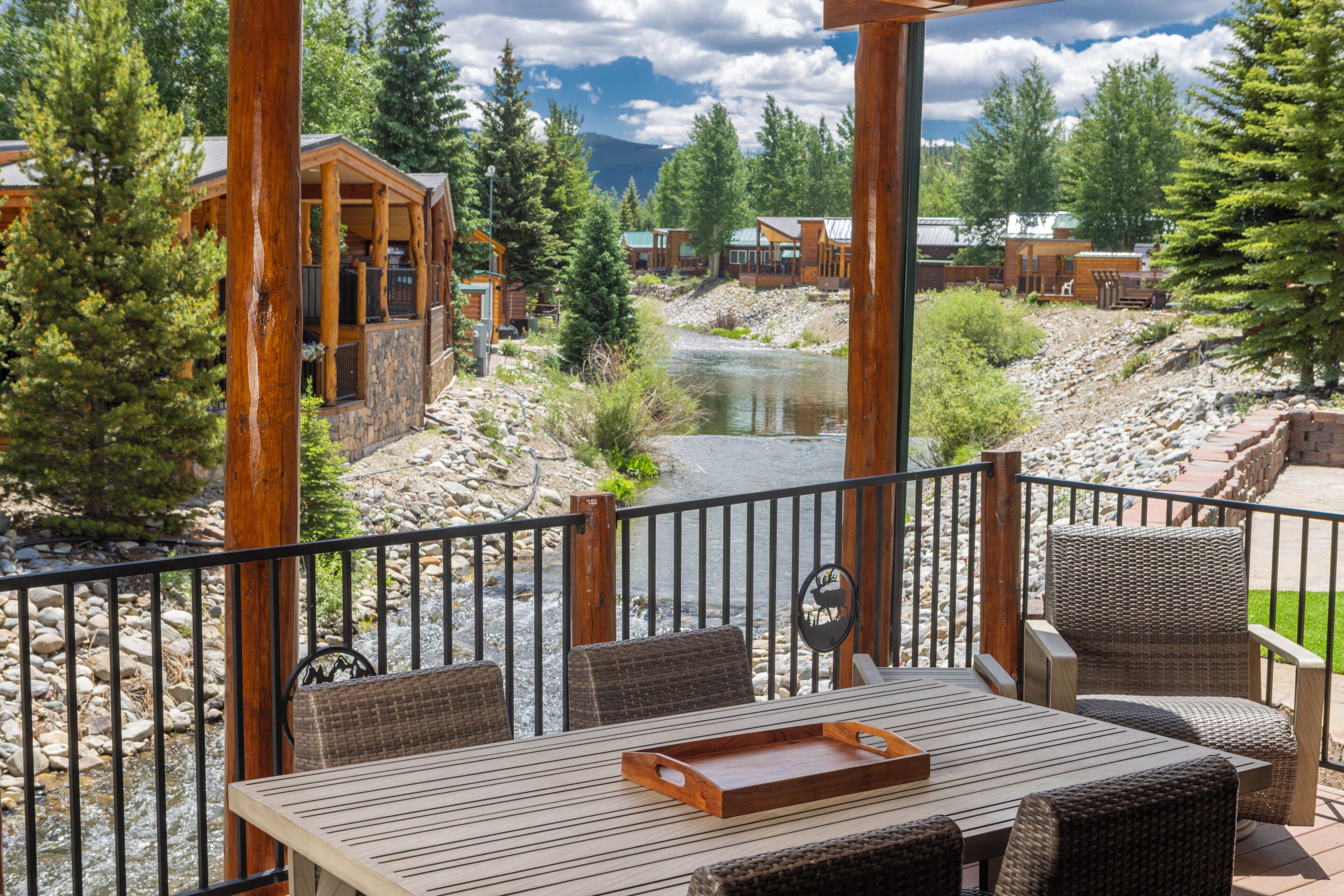 A wooden deck with outdoor furniture overlooking a river, surrounded by trees and a mountain view.
