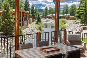 Un terrazzo in legno con mobili da esterno che si affaccia su un fiume, circondato da alberi e con vista su una montagna.