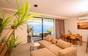 Ein modernes Wohnzimmer mit einer Couch, einem Esstisch, Stühlen und einem großen Fenster mit Blick auf das Meer.