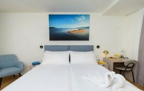 Ein Hotelzimmer mit einem Bett, einem Schreibtisch mit Lampe, einem Sessel und einem Bild mit Strandmotiv.