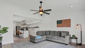 Uma sala de estar moderna com um sofá seccional, um ventilador de teto, uma bandeira e uma cozinha ao fundo.