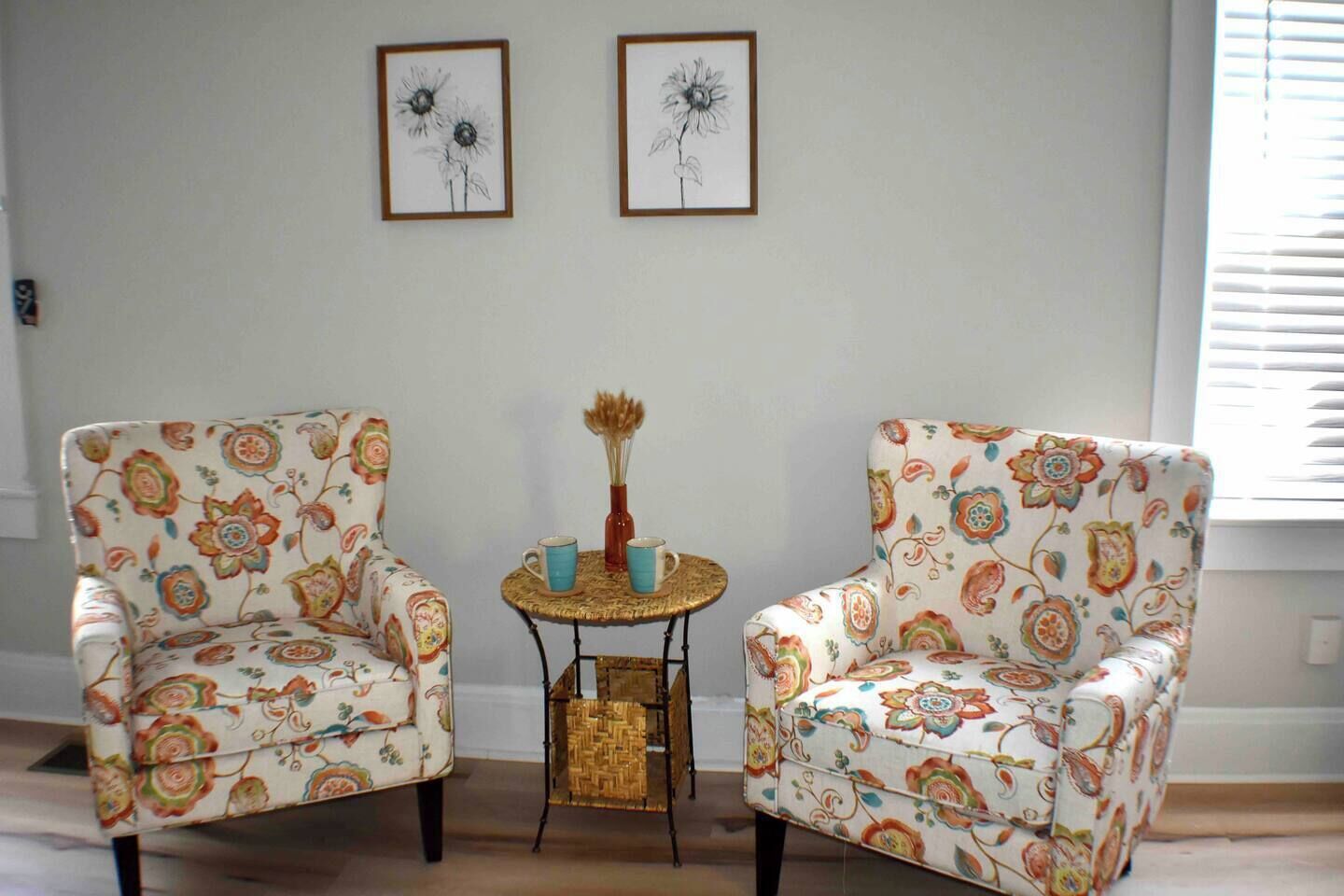 A room with two armchairs, a small round table with a vase and two cups, and two framed floral artworks on the wall.