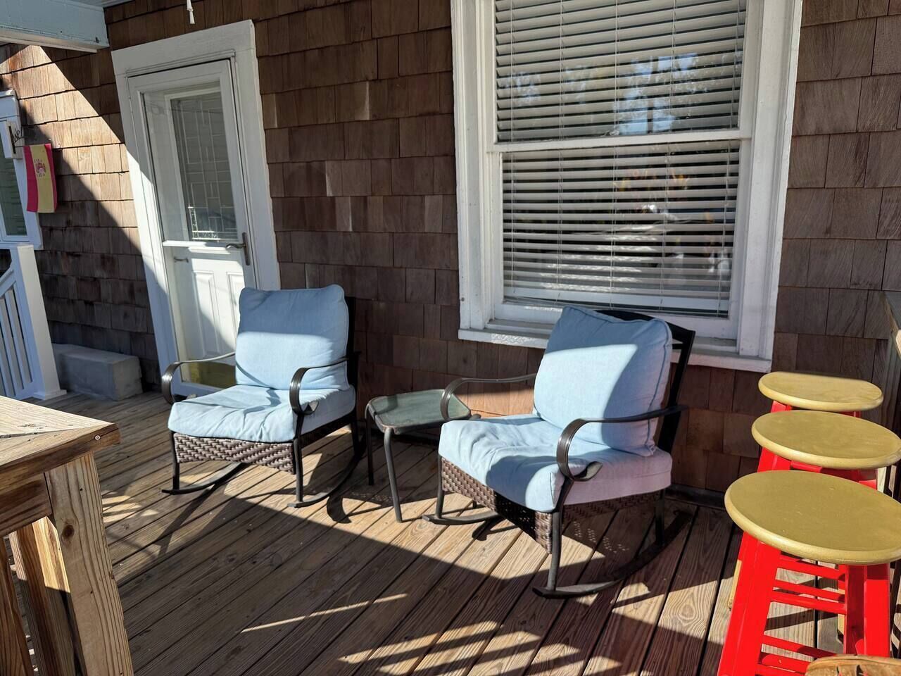 Un área de estar al aire libre con sillas de mimbre y una pequeña mesa, una terraza de madera y una ventana con persianas.