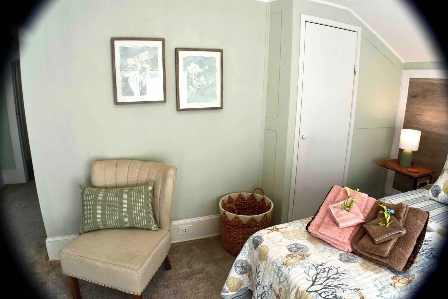 A bedroom with a chair, a bed with floral bedding, a nightstand with a lamp, and two framed pictures on the wall.