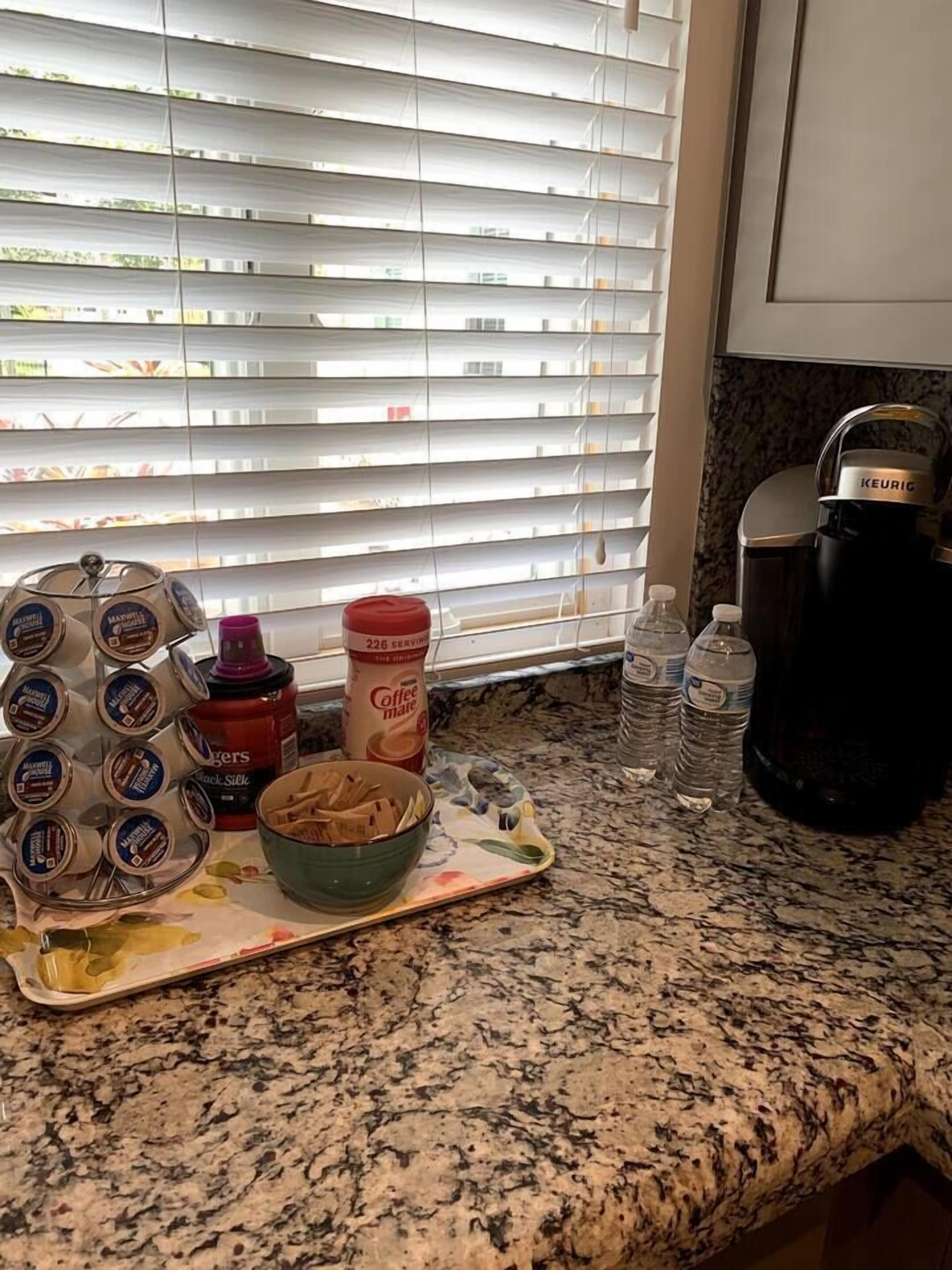 Uma bancada de cozinha com uma cafeteira, um porta-canecas, uma tigela de cereais, uma caneca de café, uma garrafa de água e um frasco de leite condensado para café.