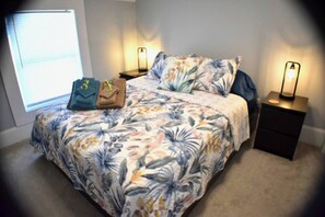 Un dormitorio con cubrecama floral, dos mesitas de noche con lámparas, una ventana con persianas y un bolso sobre la cama.