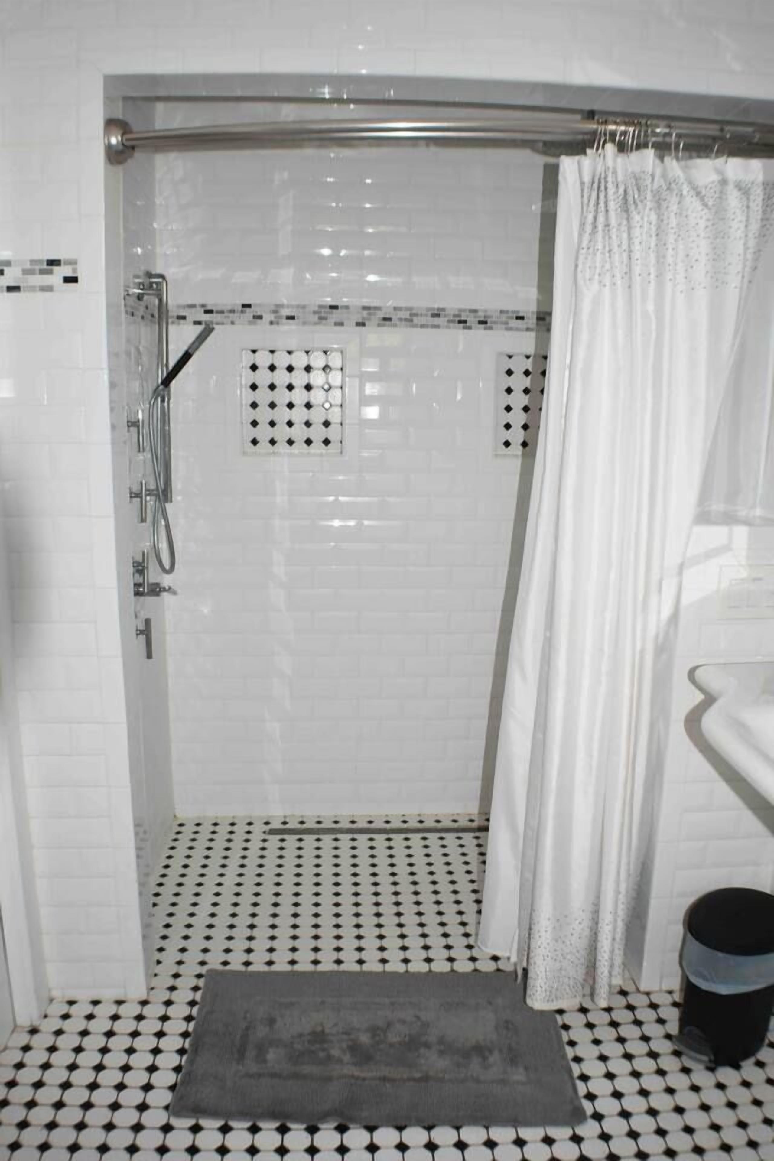 Ducha con puerta de vidrio, cortina blanca y piso estampado.