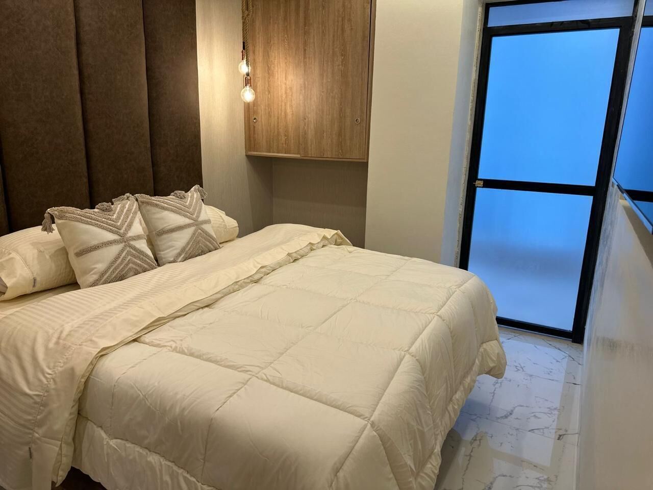 A bed with a quilted cover, a wooden headboard, and a wall-mounted light fixture.