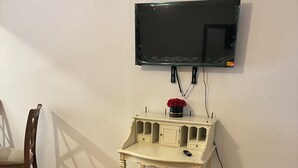 Une pièce avec un plancher en bois, un mur blanc, un téléviseur fixé au-dessus d’une petite table blanche dotée de tiroirs, et une chaise en bois.