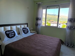 A bedroom with a bed, two pillows with motivational text, a bedside table with a lamp, and a window with a view of a landscape. - 2BR/2BA Apartment with Stunning Sunset View (Berat)