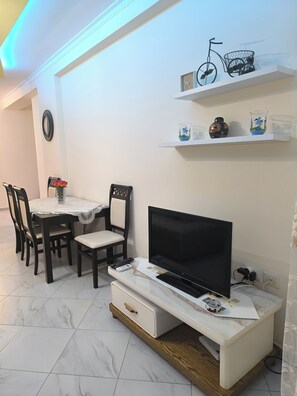Ein Wohnzimmer mit Fernseher auf einem Schrank, einem Esstisch mit Stühlen, einer Wandregal mit Dekoration und einer Uhr an der Wand.