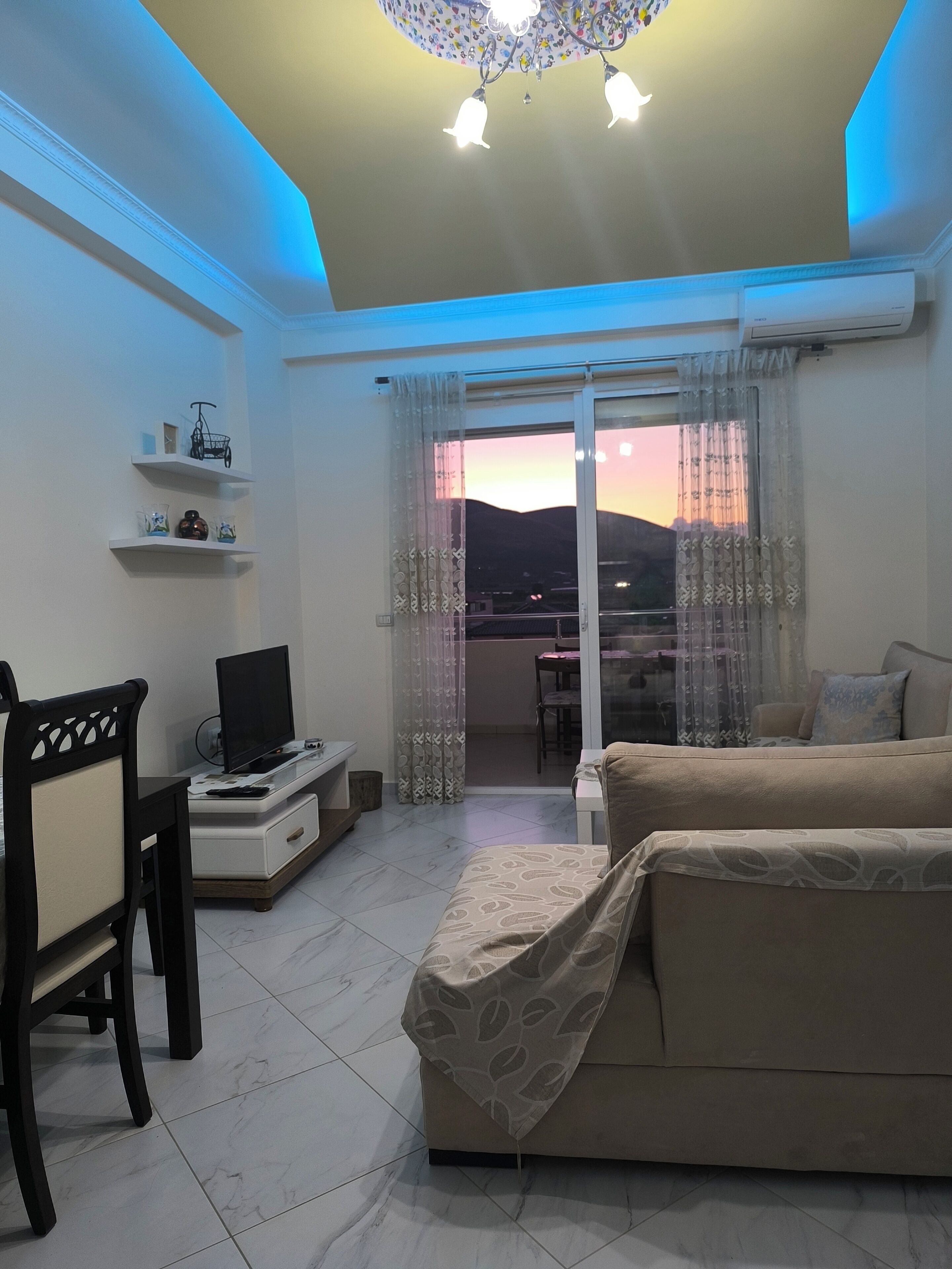 A living room with a sofa, a TV on a stand, a small table, and a chair. There is a balcony with a view of the outside.