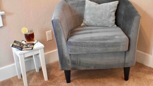 Un sillón gris con un cojín, una mesita auxiliar blanca con una bebida y un menú, y un piso cubierto de alfombra color beige.