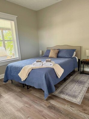 A bedroom with a bed, a nightstand, a window with blinds, and a patterned rug. - Casa Carrera 2BR/2BA Main Cottage - Walk to Town (St. Augustine)