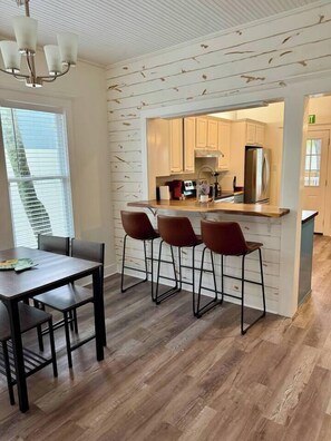 A modern kitchen with wooden paneling, a central island with stools, and a dining area with a table and chairs. - Casa Carrera 2BR/2BA Main Cottage - Walk to Town (St. Augustine)