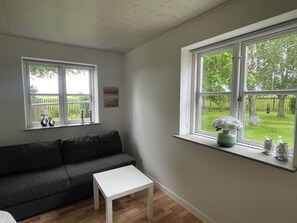 A room with a grey sofa, a white table, and a window with a view of a green garden. - Lovely big house - quiet and beautiful - with garden and fence for dogs (Horslunde)