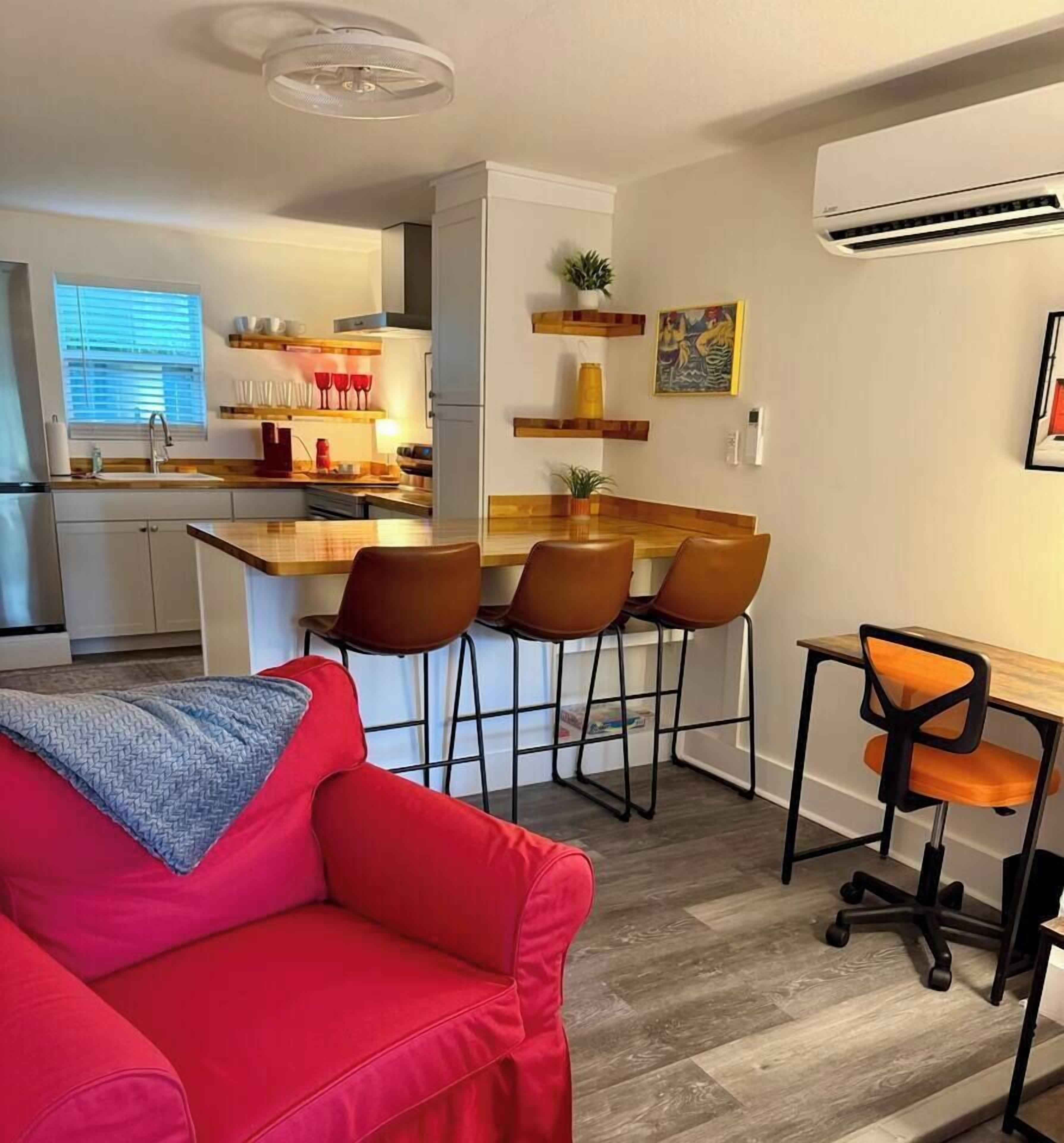 A compact living space with a kitchenette, a dining area with bar stools, a red sofa, and a small table with a chair.