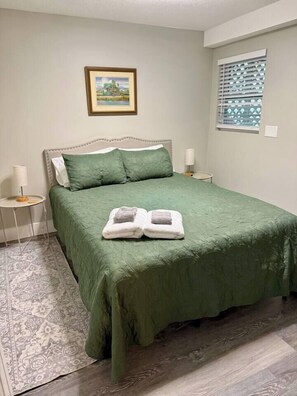 Ein Schlafzimmer mit grüner Bettdecke, zwei Kissen, einem Nachttisch mit Lampe und einem gerahmten Bild an der Wand.