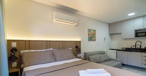 Une chambre d’hôtel comprenant un lit, une table de chevet, une lampe, un appareil de climatisation, une kitchenette et un tableau au mur.