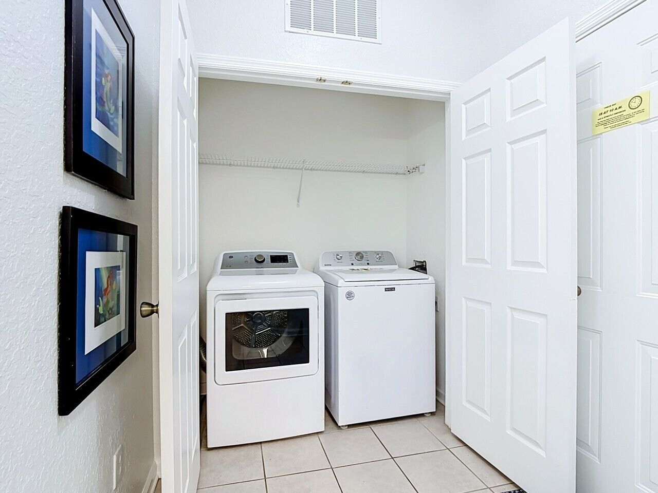 Une buanderie équipée de deux machines à laver et d’un sèche-linge, avec des murs blancs et une porte sur laquelle est apposée une pancarte.