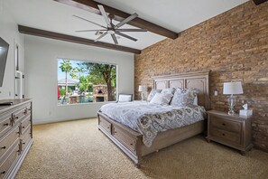 Ein Schlafzimmer mit Backsteinwand, einem großen Bett mit gemusterten Betttextilien, einem Deckenventilator und Blick auf einen Swimmingpool und Außenbereich.