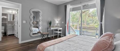 A bedroom with a bed, a desk, a chair, a mirror, and a view of a pool.