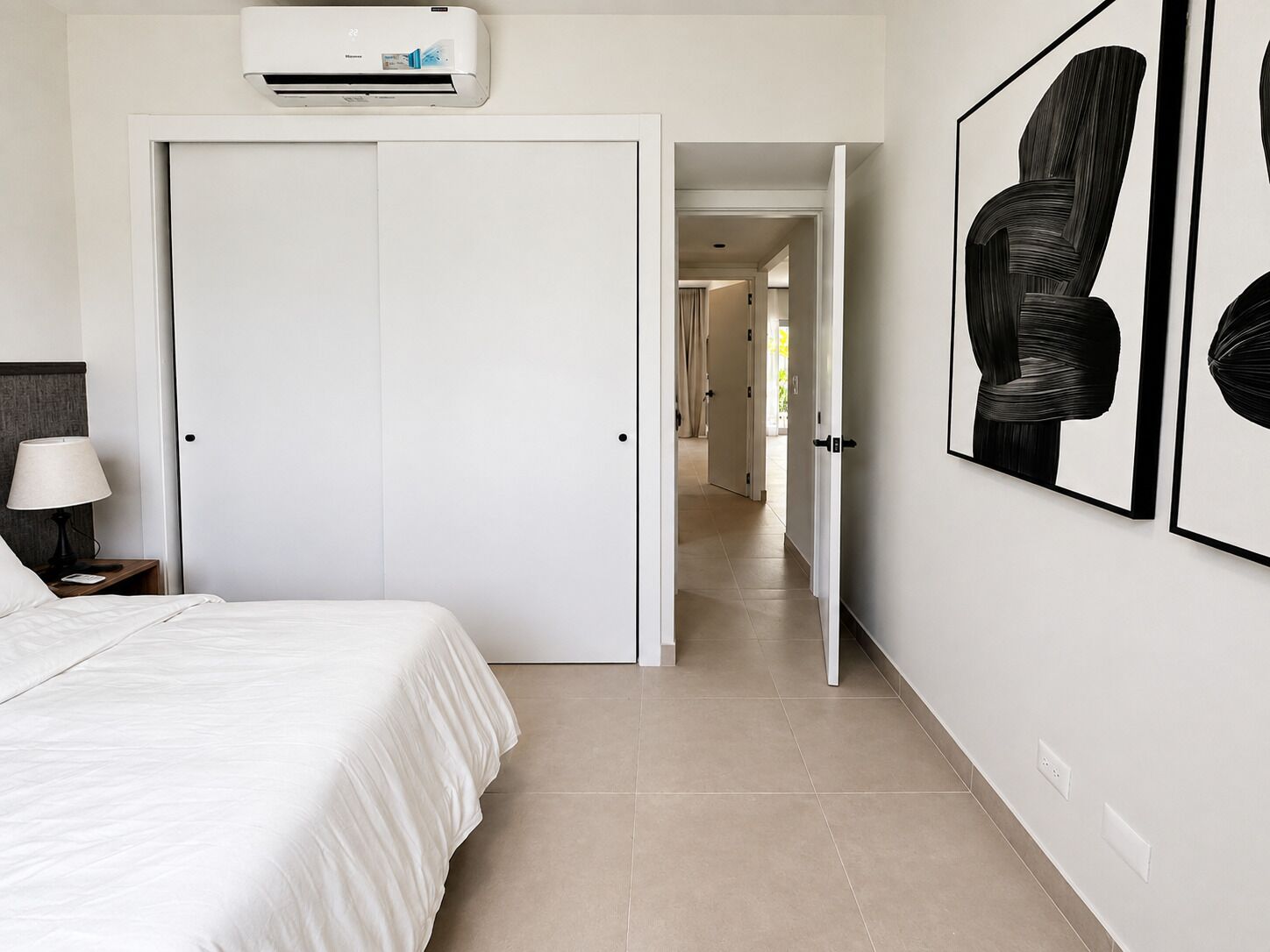 A hotel room with a bed, a nightstand, a lamp, an air conditioning unit, and a hallway leading to another room.