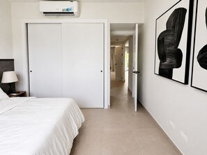 A hotel room with a bed, a nightstand, a lamp, an air conditioning unit, and a hallway leading to another room. - Poolside condo Downtown Punta Cana &$& (Punta Cana)