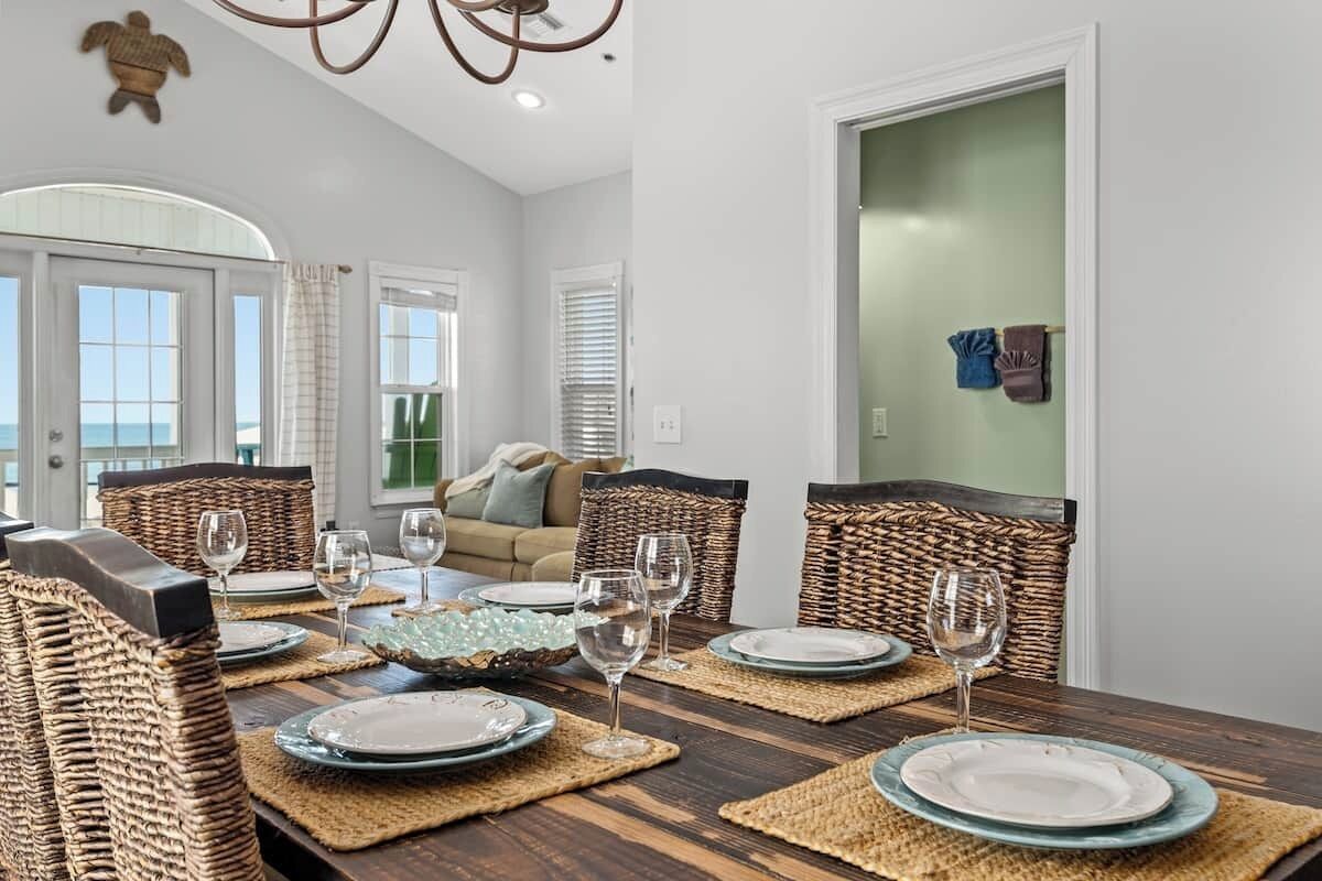 Comedor con mesa de madera dispuesta para cuatro personas, sillas de mimbre y vista al océano a través de grandes ventanales.