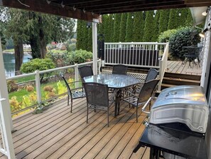 A covered outdoor patio with a round table and chairs, a grill, and a view of a lake and greenery. - Ward Lake Retreat - Lake View, Neighborhood Dock & Sports Court (Olympia)
