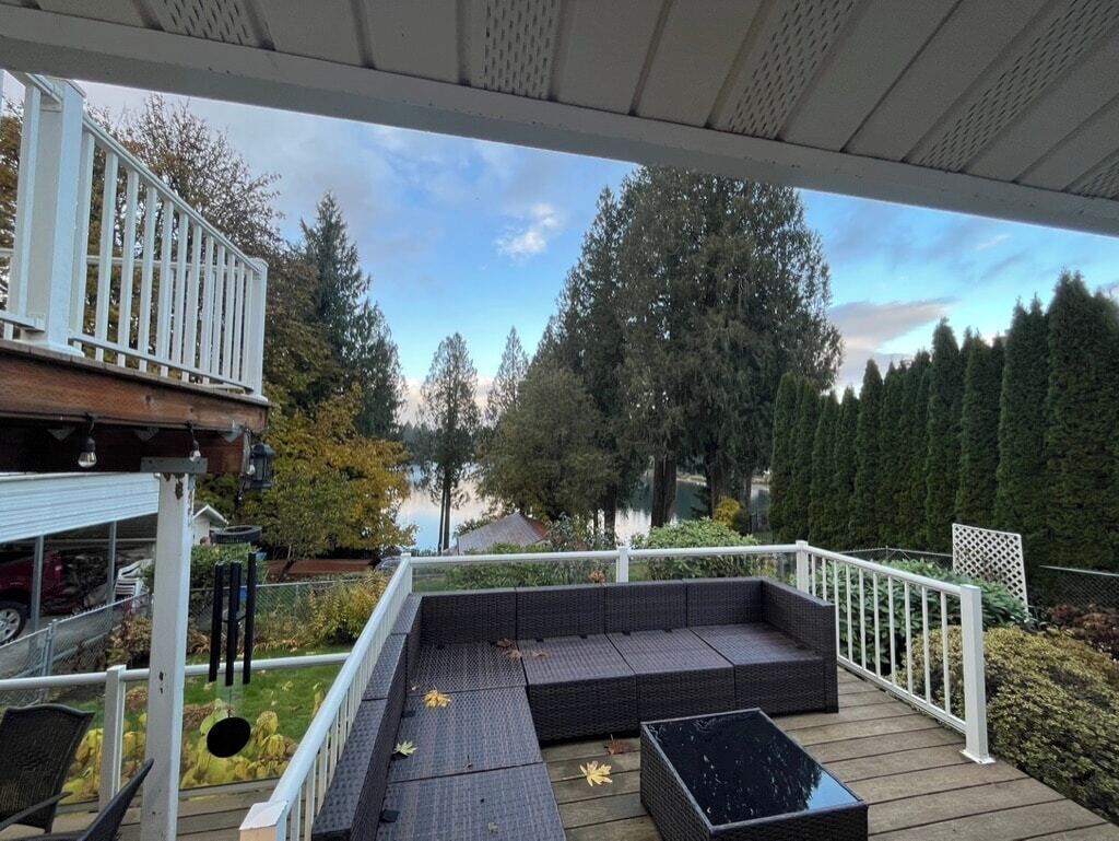 A covered outdoor deck with a dark-colored sofa and a coffee table, surrounded by a white railing and a view of trees and a body of water.