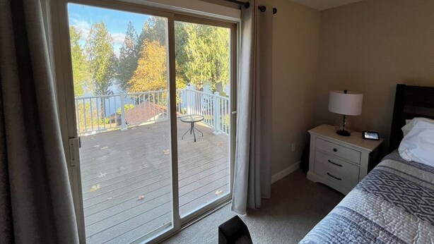 A hotel room with a large sliding glass door leading to a balcony, a bed with a patterned bedspread, a bedside table with a lamp, and a small table on the balcony. - Ward Lake Retreat - Lake View, Neighborhood Dock & Sports Court (Olympia)