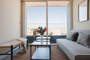 A modern living room with a sofa, a coffee table, and a bench. A large window offers a view of a cityscape. - Apartment Nearby Metro with Pool (Ñuñoa)