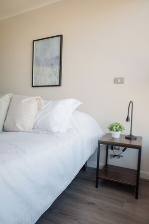 A bedroom with a bed, a nightstand with a lamp, a small plant, and a painting on the wall. - Apartment Nearby Metro with Pool (Ñuñoa)
